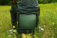 Person sitzt auf altem Röhrenfernseher in Blumenwiese