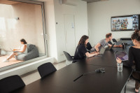 Menschen sitzen im Büroraum, auf dem Bildschirm sind Kacheln von Menschen zu sehen, die digital zugeschaltet sind. Draußen sitzt eine Mitarbeiterin auf einem Sitzsack vor ihrem Laptop.